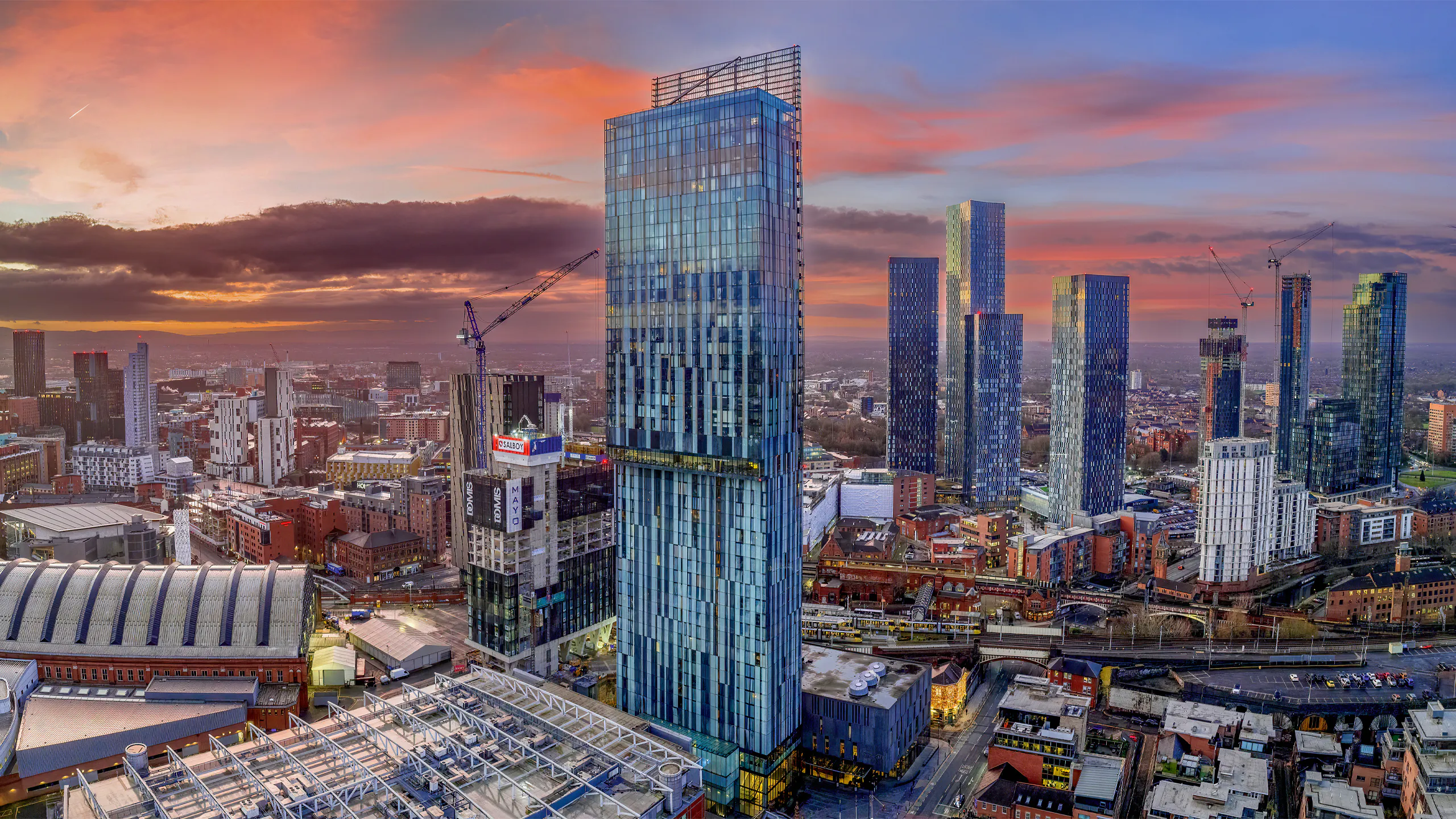 Manchester cityscape with high-rise buildings and construction cranes under a dramatic sunset sky.
